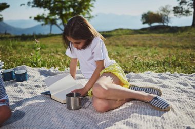 Mutlu bir çocuk yaz pikniği sırasında açık havada kitap okuyor. Doğayla iç içe, çarpıcı bir dağ ortamında, sıcak ve güneşli bir günün tadını çıkarıyor..