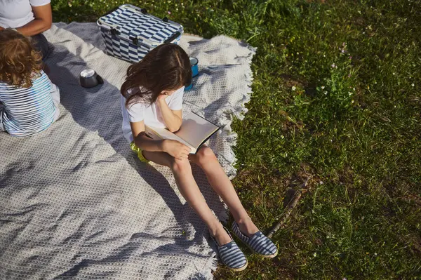 Bir aile, yeşilliklerle çevrili bir battaniyenin üzerinde huzurlu bir açık hava pikniğinin tadını çıkarıyor. Çocuklar kişisel aktivitelerle uğraşırken birlikte geçirdikleri zamanın tadını çıkarırken ortam sakin..