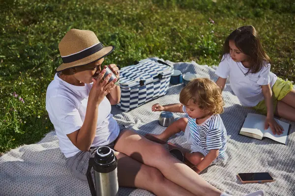 Sevgi dolu bir anne ve iki çocuk yemyeşil ortamda huzurlu bir pikniğin tadını çıkarıyorlar. Çocuklar nişanlı ve mutlular, beraberliğin özünü ve açık hava aktivitelerinin neşesini yakalıyorlar..