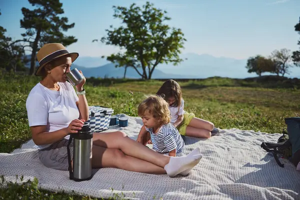 Anne ve çocuklar dağlarda piknik yaparken dışarıda kaynaşıyorlar. Görüntü, güzel bir doğal ortamda aile birliği, rahatlama ve doğanın tadını çıkarıyor..