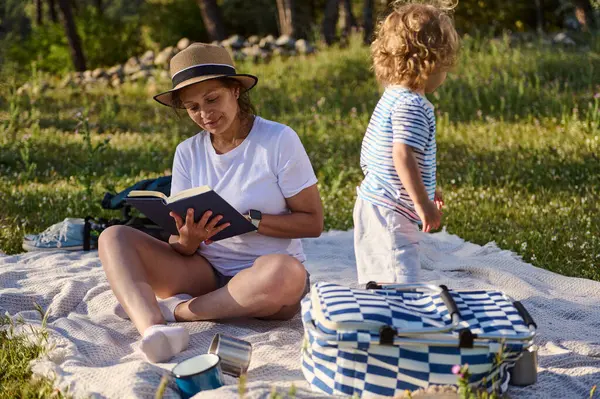 Bir anne çocuğuyla battaniyeye oturmuş dışarıda eğlenirken kitap okur. Sahne sakinlik, bağ kurma ve gevşeme sergiliyor manzaranın manzarası.