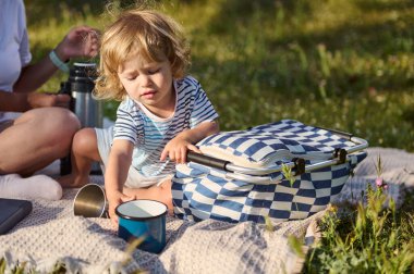 Küçük bir çocuk açık havada piknik yapmaktan hoşlanıyor, bir sepetin yanındaki battaniyeyle meşgul, güneşli gökyüzünün altında çimenli yeşilliklerle çevrili, boş zamanlarında keyifli bir anı paylaşıyor..
