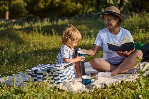 Bir anne, çocuğuyla birlikte yeşil bir çevrede piknik yaparken, sıcak içecekler okurken ve paylaşırken, mutluluk ve neşe anlarını paylaşırken, manzaralı bir ortamda kaliteli zaman geçirmekten zevk alır..