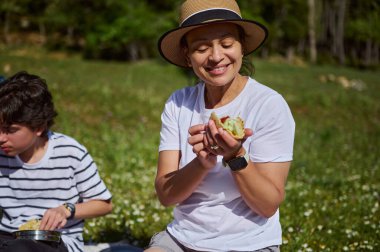 Doğal bir dağda aile pikniğinde taco yerken gülümseyen bir kadın. Neşeli bir bağ anı, doğa ve paylaşılan bir yemek keyfi..