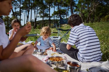 Bir anne ve çocukları, bereketli yeşillik, beraberlik ve sağlıklı bir yaşam tarzıyla çevrili iken, huzurlu bir açık hava ortamında keyifli bir pikniği paylaşıyorlar..