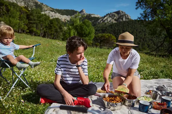 Bir anne ve çocukları manzaralı bir dağ manzarasının ortasında lezzetli tacoların tadını çıkarırlar. Neşeli an beraberliği, açık hava eğlencesini ve doğanın güzelliğini vurgular..
