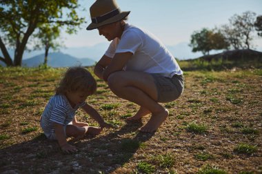 Bir anne, ağaçlarla çevrili çimenli bir arazide, doğadaki aile bağlarını ve keşiflerini ön plana çıkararak yavrusunun yanına çömelir..
