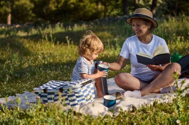 Bir anne, çocuğuyla birlikte yeşil bir çevrede piknik yaparken, sıcak içecekler okurken ve paylaşırken, mutluluk ve neşe anlarını paylaşırken, manzaralı bir ortamda kaliteli zaman geçirmekten zevk alır..