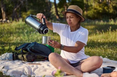 Güneşli bir ormanda sakin bir piknik sırasında termostan su döken bir kadın. Konforlu ortam, sıcak ışık ve sakin ortam rahatlama ve doğa takdirini iletir..