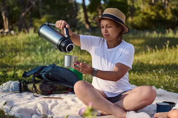 Güneşli bir ormanda sakin bir piknik sırasında termostan su döken bir kadın. Konforlu ortam, sıcak ışık ve sakin ortam rahatlama ve doğa takdirini iletir..