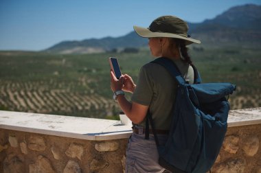 Sırt çantalı, doğa dolu bir ortamda tek bir kişi akıllı telefona bakıyor. Sahne, resmedilmeye değer dağlık bir ortamda sakin bir seyahat ve keşfi vurguluyor..