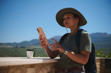 Bir gezginin açık hava sahnesinde şapka ve sırt çantasıyla gezerken yanında kahve ve akıllı bir telefonla manzaralı bir dağ manzarasına karşı..
