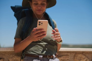 Sırt çantası ve şapkası olan bir gezgin açık havada, açık havada, açık havada, mavi gökyüzünün altında telefonlarını kontrol ediyor. Macera ve bağlantıyı temsil etmek.