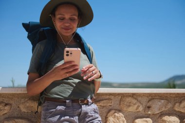 Sırt çantalı ve şapkalı kadın akıllı telefonunu kontrol ederken macera yolculuğu sırasında manzaralı bir yeri keşfediyor..