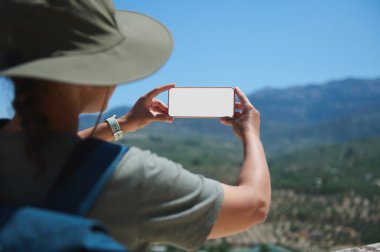 Güneşli bir günde göz kamaştırıcı dağ manzaraları yakalamak için akıllı telefon kullanan şapka takan bir turist doğayı ve seyahat keşiflerini gözler önüne seriyor..