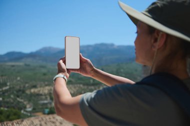 Akıllı telefonuyla parlak, güneşli bir arkaplan karşısında resimli bir dağ manzarası yakalayan biri. Görüntü, keşifleri, doğanın neşesini ve teknolojinin keşiflere entegrasyonunu iletir..