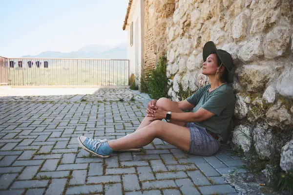Kırsal taştan bir duvarın yanında oturan ve arka planda geniş dağ manzaraları olan sakin bir çevrenin tadını çıkaran bir kişinin sakin bir sahnesi. Barışçıl açık hava seyahatinin mükemmel bir temsili..