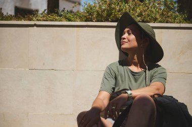 Açık havada oturan bir kadın geniş yeşil bir şapka ve haki rengi elbiseler giyerek güneşli bir günün tadını çıkarıyor. Duvar ve yeşil bitkiler huzur ve rahatlığı somutlaştıran huzurlu ve doğal bir atmosfer yaratırlar..