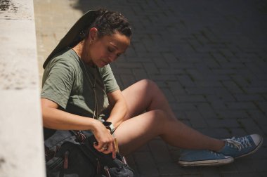 Genç bir kadın, rahatlık ve keşif için giyinmiş, güneşli havada, rahat ve kentsel bir yaşam tarzını yansıtan çantasıyla rahatça oturuyor..