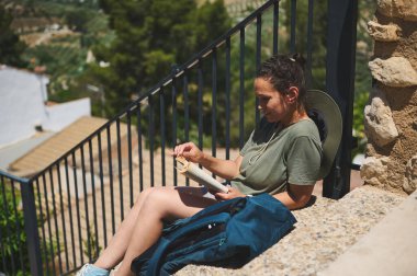 Bir gezgin açık havada güneşin altında oturmuş resimli kırsal ve dağlık bir manzarayla çevrili bir kitabın tadını çıkarıyordu. Görüntü sakinliği, rahatlığı, rahatlığı ve doğayla bağlantıyı iletir..