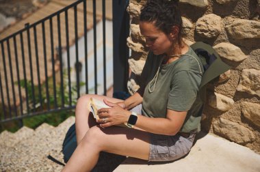 Bir kadın, tarihi bir dağ manzarasında aktif turizm ve doğa takdirini temsil eden ortaçağ taşlarından oluşan manzaralı bir zemin karşısında kitap okuyarak huzurlu bir anın tadını çıkarıyor..