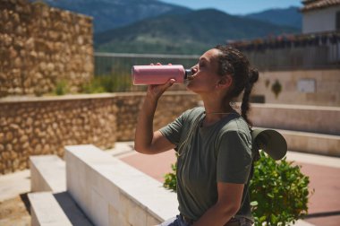Genç bir kadın susuzluğunu suyla giderirken dağlarla çevrili bir ortaçağ şehrinin manzarasının tadını çıkarıyor. Aktif seyahat ve açık hava keşiflerini temsil ediyor..