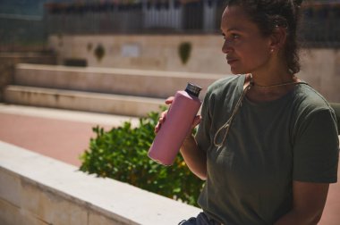 Genç bir kadın elinde pembe bir şişeyle bankta dinleniyor. Güneşin altında manzaralı bir günün tadını çıkarıyor. Rahatlama ve farkındalık taşıyor..