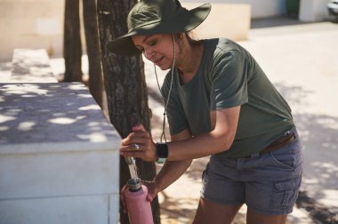 Bir kadın yaz güneşinin altında açık havuzda biberonunu dolduruyor. Geniş kenarlı bir şapka takıyor, sıcak bir günde hazırlığını ve sürdürülebilirliğini gösteriyor. Ekolojik bilinç yolculuğunun mükemmel bir temsili..