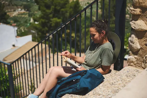 Bir gezgin açık havada güneşin altında oturmuş resimli kırsal ve dağlık bir manzarayla çevrili bir kitabın tadını çıkarıyordu. Görüntü sakinliği, rahatlığı, rahatlığı ve doğayla bağlantıyı iletir..