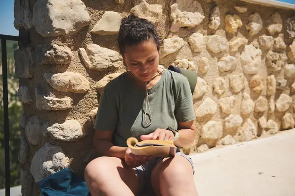 Bir kadın, gün ışığında otururken bir kitabı inceler taş duvarların manzaralı, kırsal arka planıyla, doğa ve tarihi harmanlayarak açık hava ortamında..