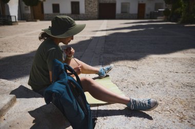 Sıradan giyinmiş bir insan bir meydanda oturmuş haritayı analiz ediyor ve Akdeniz atmosferiyle manzaralı bir ortam sergiliyor..