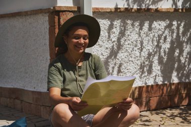 Neşeli bir gezgin güneşli bir bölgede açık hava haritası okumaktan zevk alır, rahat bir şapka ve günlük giysiler giyer. Arka planda duvarlar boyunca gölgelerin oynadığı rustik mimari sergileniyor.