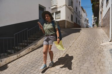 Bir maceraperest harita ve akıllı telefon kullanarak güzel bir İspanyol köyünde, kaldırım taşı caddesinde geziniyor. Güneşli gün ve huzurlu ortam, tarihi çevrenin güzelliğini vurguluyor..