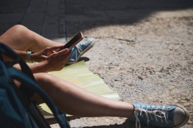 Bir gezgin dışarıda akıllı bir telefonla oturuyor, yerde bir harita ve günlük kıyafetlerle, güneşli bir yerde keşif ve planlama öneriyor..