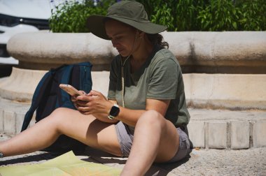 Bir insan, güneşli bir günde seyahat ve keşfin özünü yakalayan büyüleyici bir ortamın arka planına karşı, akıllı bir telefon ve sırt çantasıyla dışarıda dinlenir ve navigasyon araştırması yapar..