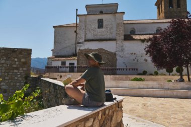 Bir gezgin, bir İspanyol köyünde antik bir binanın yanında, elinde bir harita tutarak ve yakınlardaki dağların manzarasının tadını çıkararak, macera ve keşifleri ön plana çıkarıyor..