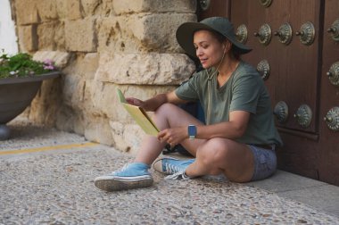 Bir gezgin tarihi bir binanın dışında oturmuş, rahat ve güneşli bir atmosferde harita üzerinde çalışıyor. Ortaçağ mimarisi ve manzara çevresi macera ve kültürel keşfi çağrıştırıyor..