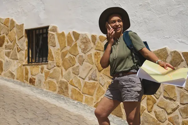 Bir gezgin telefonda konuşurken elinde bir harita tutarak tarihi bir köyde geziniyor. Kaldırım yolu ve kırsal taş duvarlar manzaralı bir atmosfere ekleniyor..