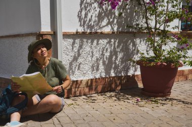 Bir gezgin elinde harita tutarak bir Akdeniz köyünde huzurlu bir anın tadını çıkarıyor. Sahne keşif, doğa ve güneşli bir günü betimliyor. Saksı çiçekleri ve gölgeler dinginliği arttırır..