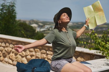 Dışarıda gülümseyen bir kadın, elinde parlak bir güneş ışığıyla bir harita tutuyor. Yeşil ve dağlık ufuk çizgisi ile çevrili taşlık bir avluda keşif ve neşe hissi..