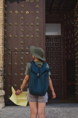 Macera teçhizatı taşıyan bir turist elinde harita olan tarihi bir İspanyol ortaçağ şehrini keşfediyor. Büyük ahşap kapı ve karmaşık taş işçiliği mekanın kültürel önemini vurguluyor..