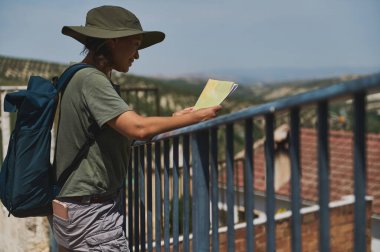 Şapkalı bir turist, açık havada, güneşli bir günde tarihi bir İspanyol köyünün ve uzak dağların manzaralı güzelliğinin tadını çıkarırken, macera ve keşif çağrıştıran bir harita tutuyor..