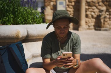 Açık havada oturan rahat bir gezgin, akıllı telefonuna dalmış. Bir şapka takarak, kısmen tarihi bir taş yapının gölgesinde kalırken sıcak havanın tadını çıkarıyor..