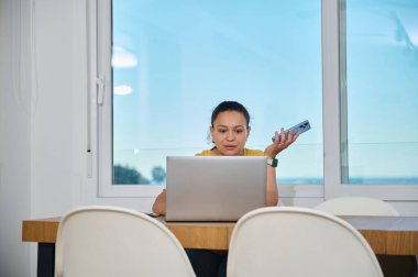 Profesyonel bir kadın ev ofisinden bilgisayar ve akıllı telefon kullanarak görevlerini yürütüyor. Sahne, üretkenliği ve adaptasyonu rahat ve modern bir mekanda yakalıyor..