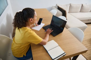 Bilgisayarlı ve defterli bir kadın ahşap bir masada ev ofisinden çalışıyor. Konsantrasyon, verimlilik ve konforlu çalışma ortamı gösteriyor. Modern uzak çalışma yaşam tarzını simgeliyor..