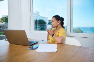 Parlak bir ev ofisteki bir kadın, etrafı temiz bir düzenek ve sahil manzarasıyla çevrili bir şekilde dizüstü bilgisayarıyla çalışırken kahve molası veriyor. Çalışmanın ve rahatlamanın ilham verici bir karışımı..