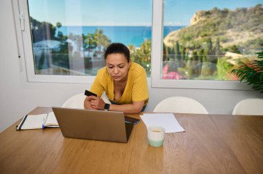 Parlak bir yaşam alanından dizüstü bilgisayarı üzerinde çalışan bir kadın. İlham verici bir kıyı manzarası var. Bağımsız yaşam tarzını somutlaştırıyor. İfadeler odak, serbest meslek ve dijital göçebe özlemleri.