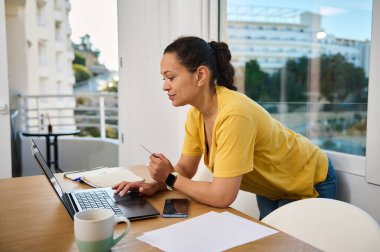 Sarı tişörtlü bir kadın, kapalı bir ortamda bir masada dizüstü bilgisayarla çalışıyor. Etrafını saran kahve fincanı, telefon ve belgeler verimli bir çalışma ortamı olduğunu gösteriyor..