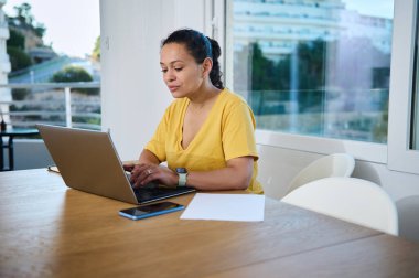 Odaklanmış sarı tişörtlü bir kadın parlak bir ofiste otururken laptopuyla dikkatlice çalışıyor. Uzak çalışma, üretkenlik ve girişimciliği temsil eder.