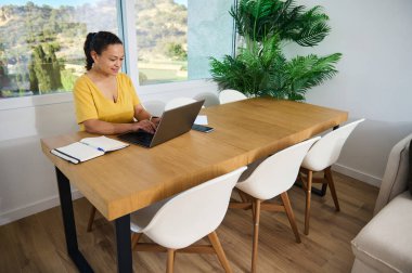 Şık bir ev ofisinde dizüstü bilgisayarda çalışan kendine güvenen bir kadın üretkenliği, girişimciliği ve doğal çevreyle çevrili rahat bir çalışma alanını vurguluyor..